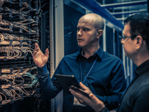Two men stand in front of an open computer server analyzing it's components while holding a tablet.